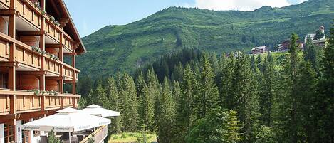 A wooden chalet with balconies and outdoor seating, surrounded by lush green mountains and forest.