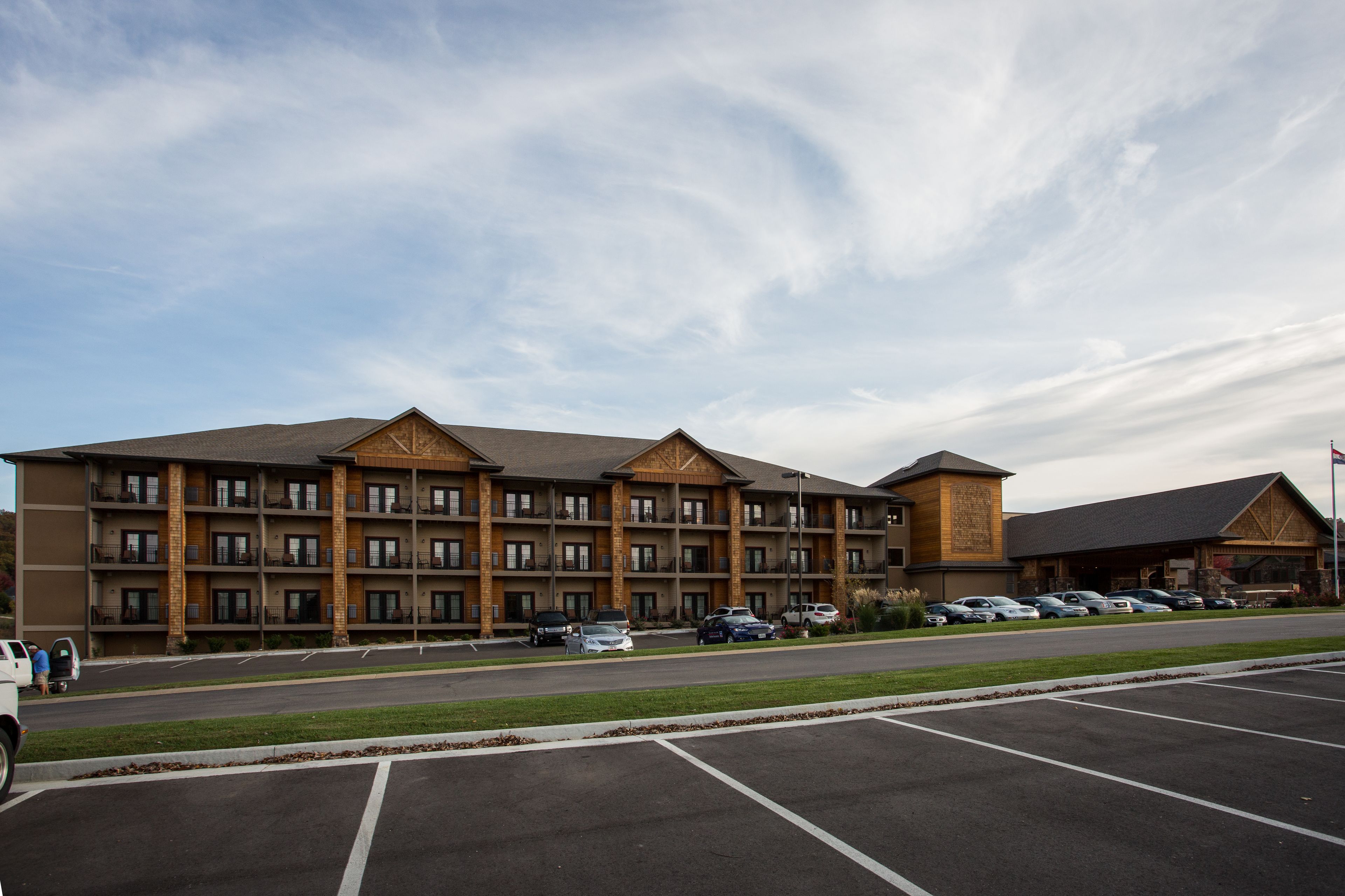 A multi-story hotel with a brick facade, multiple balconies, and a parking lot with several cars.