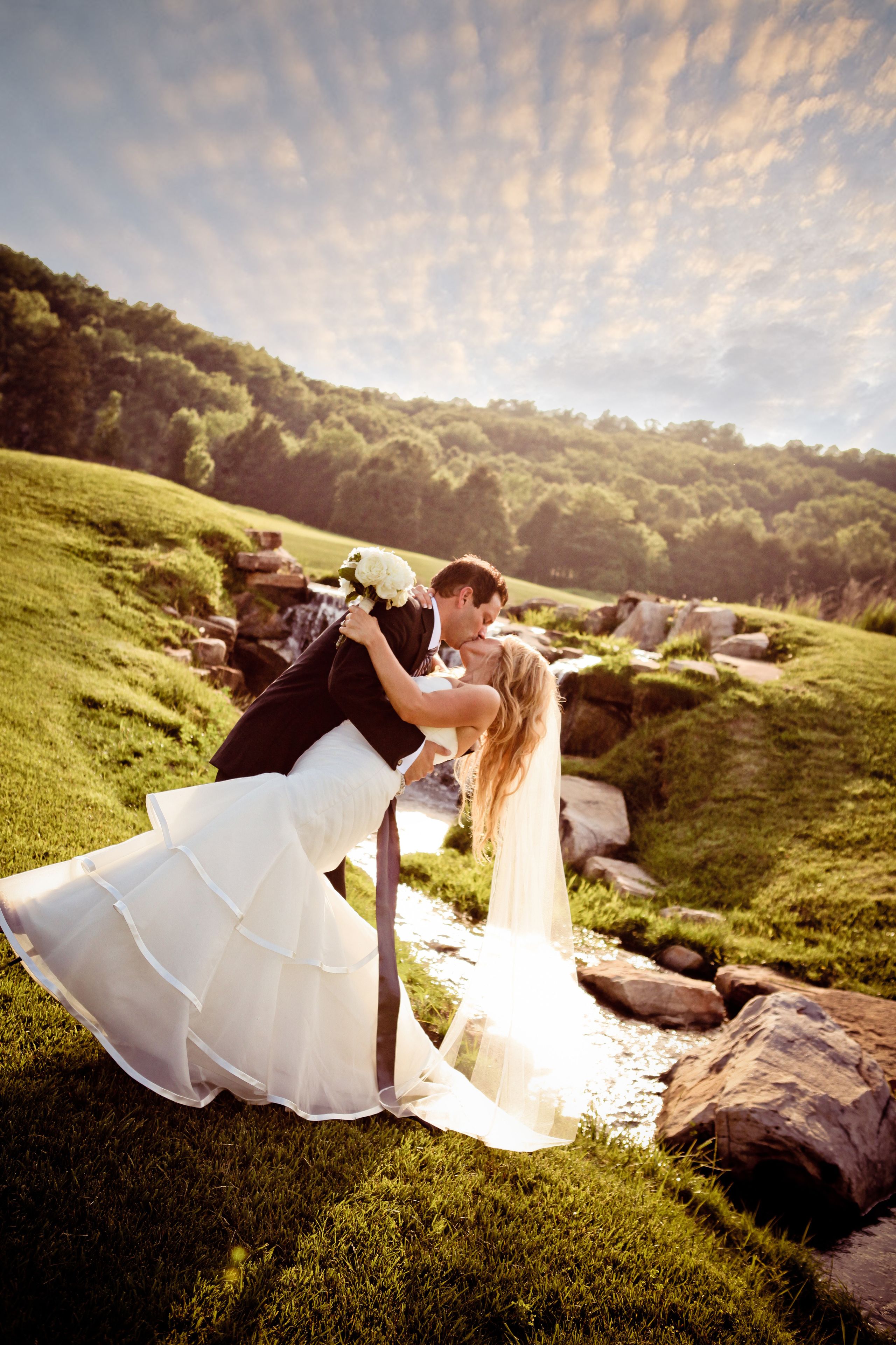 A couple embracing in a grassy outdoor setting with a stream and rocks.