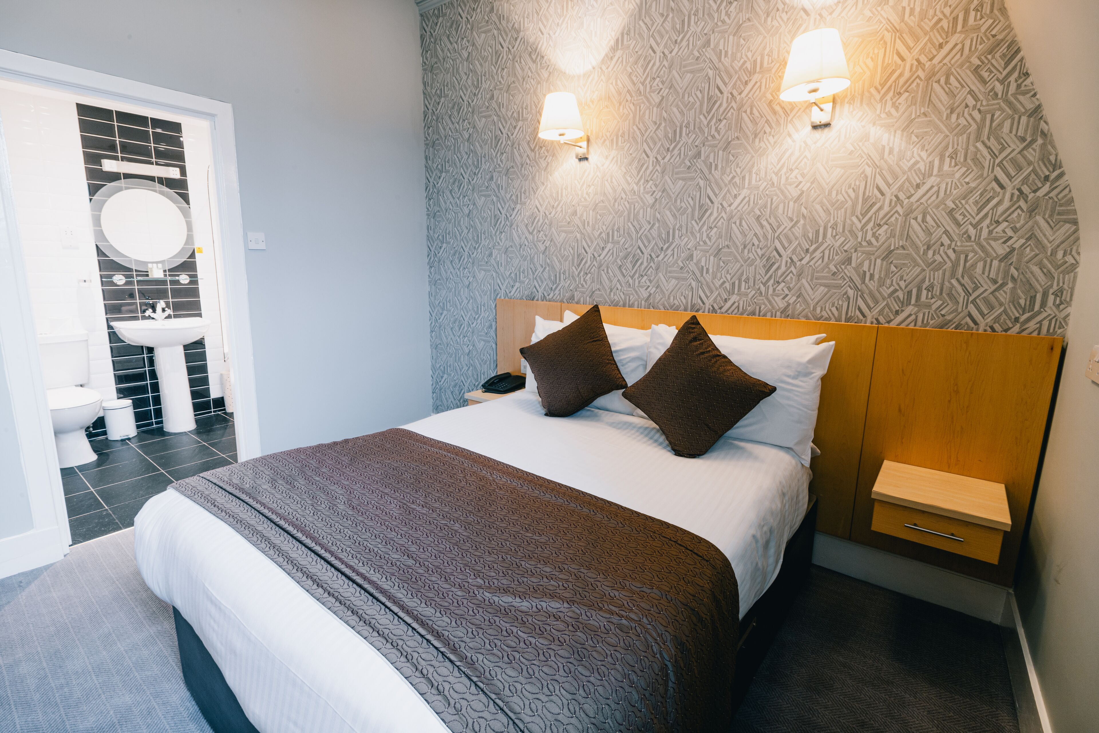 A hotel room with a bed, bedside table, nightstand, and a bathroom visible through an open door.