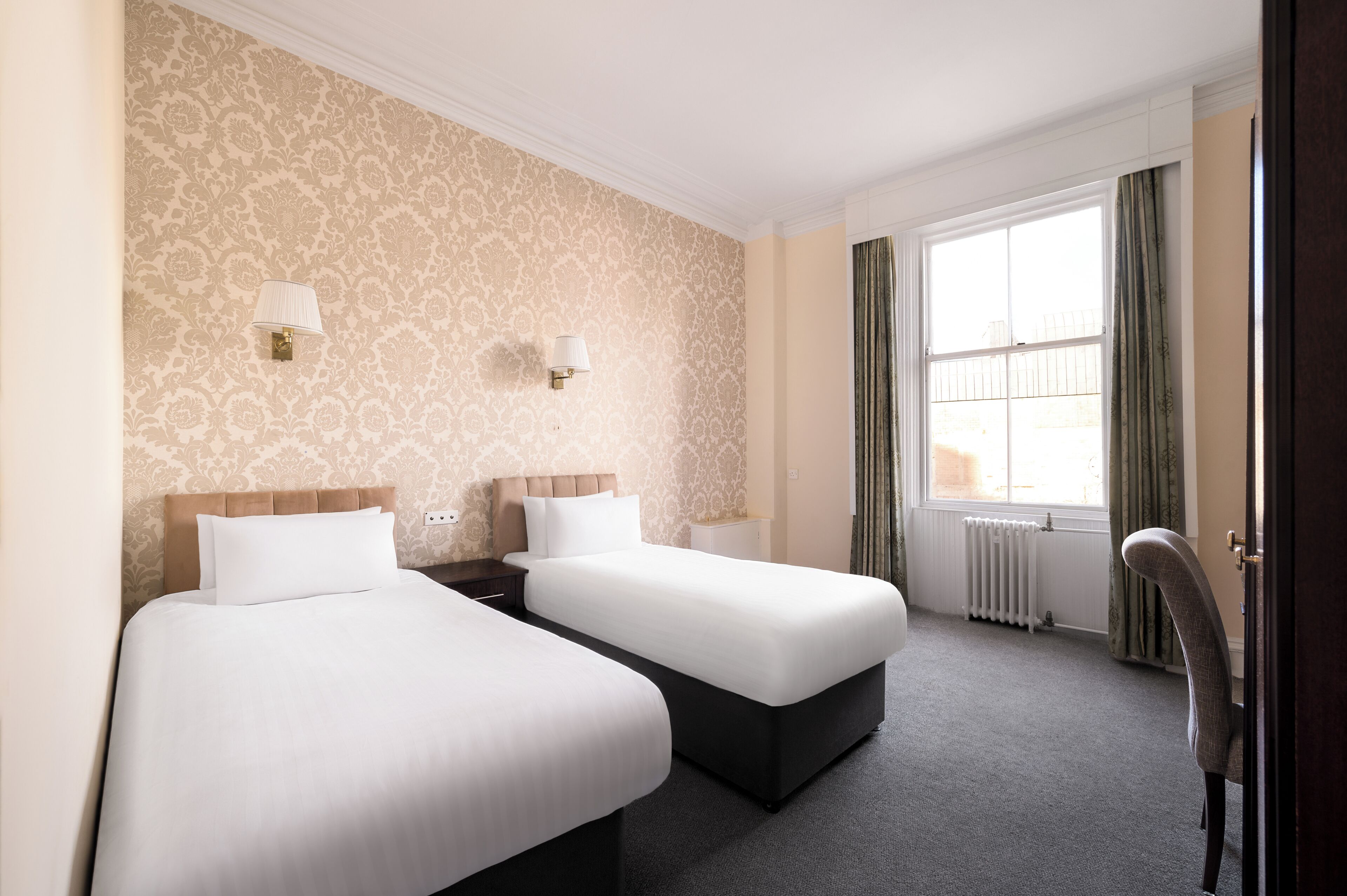 A hotel room with two beds, a window with curtains, a radiator, and a chair.