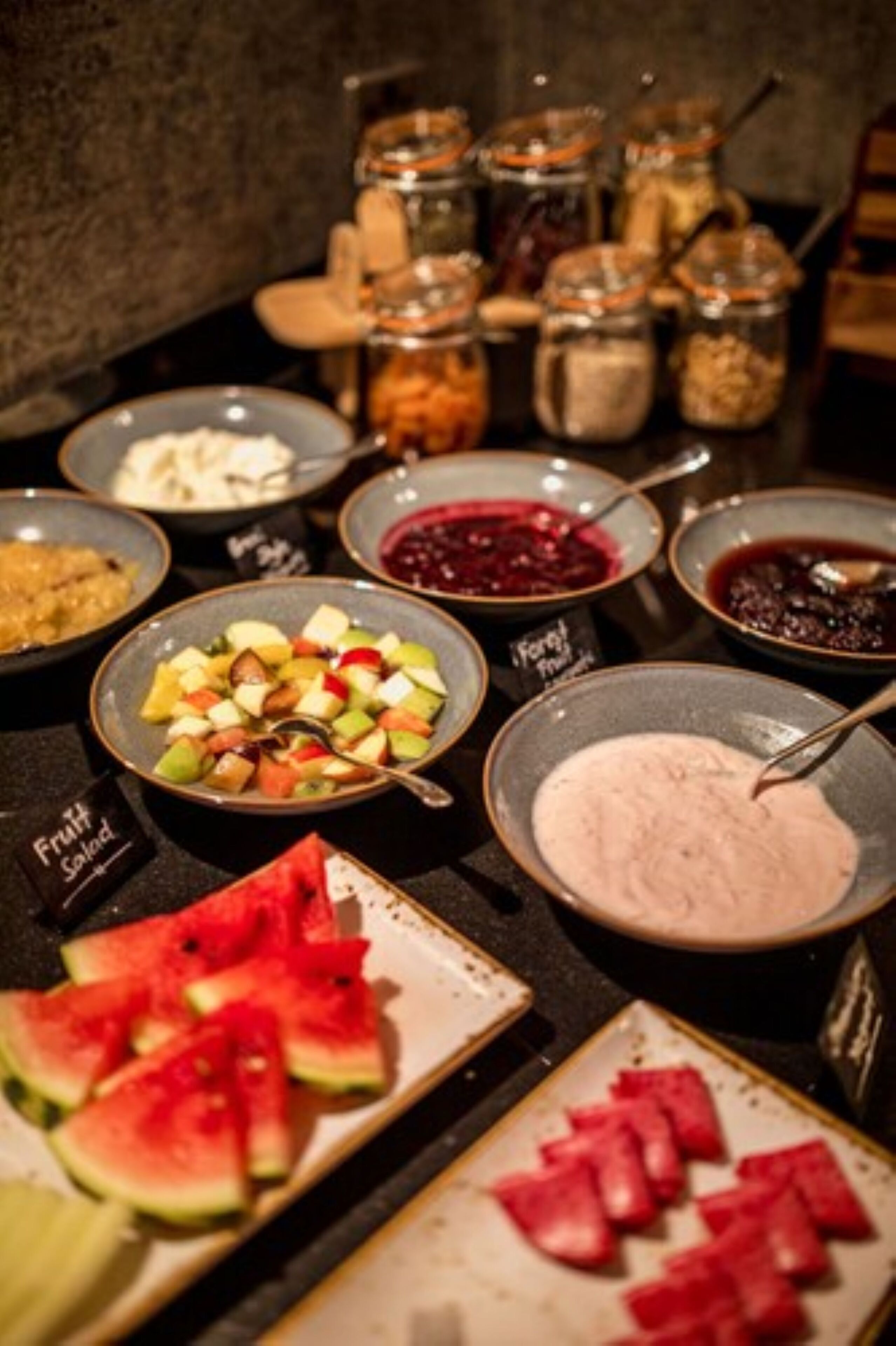 A buffet with various food items including fruit salad, watermelon slices, and different types of sauces.