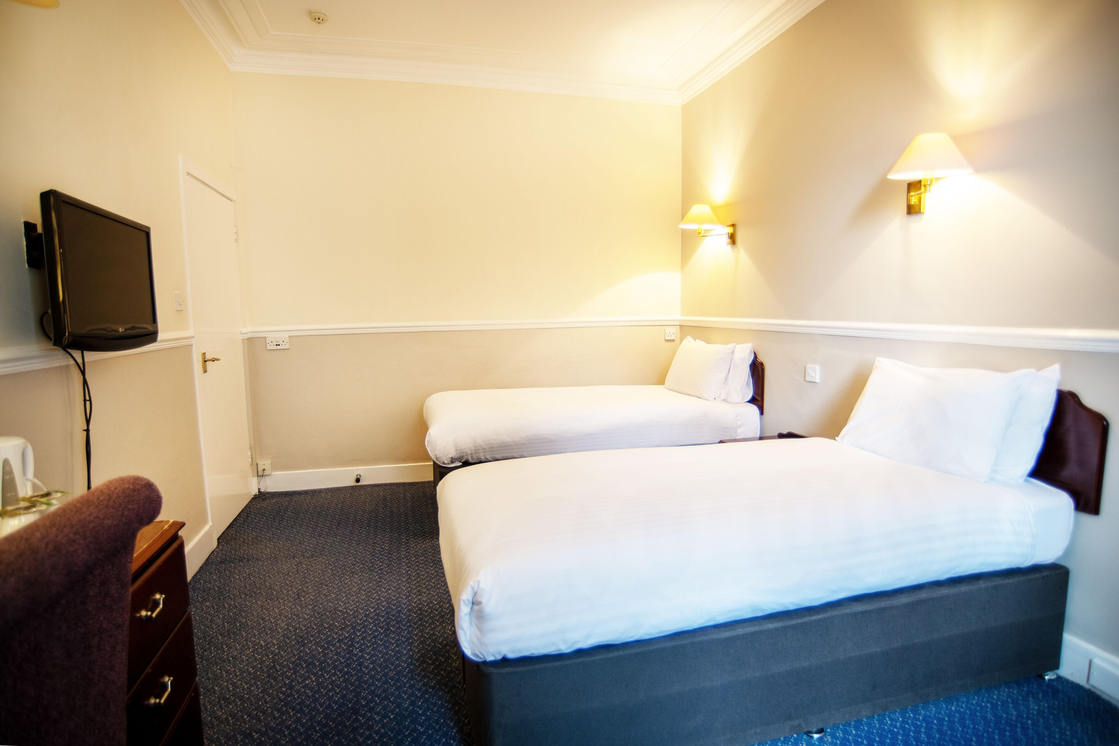 Une chambre d’hôtel avec deux lits simples, un téléviseur à écran plat, un bureau avec une chaise et deux lampes fixées au mur.