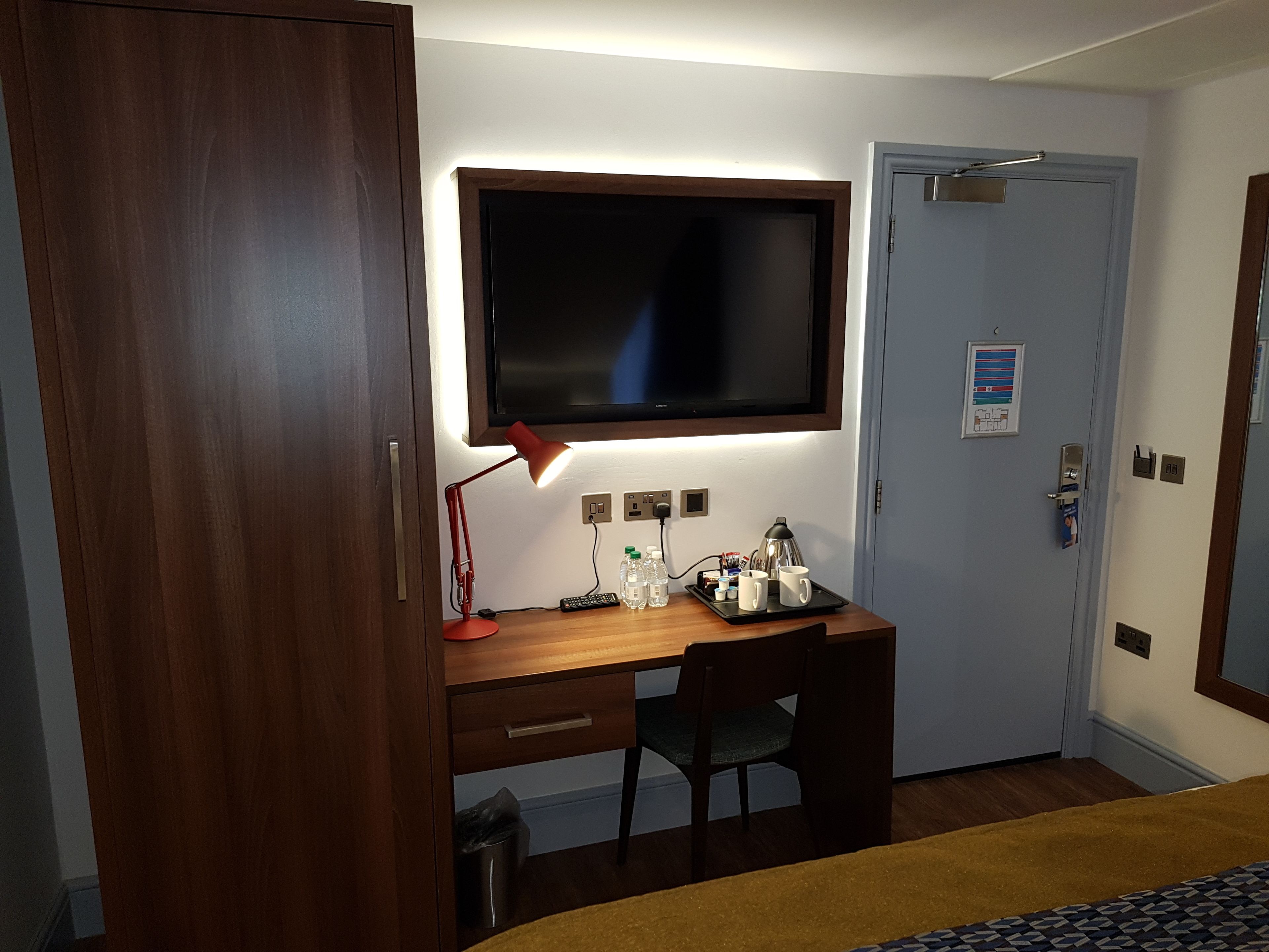Una camera d'albergo con una scrivania in legno, una sedia, una TV montata sopra la scrivania, un comodino con una lampada e un letto con copriletto decorato.
