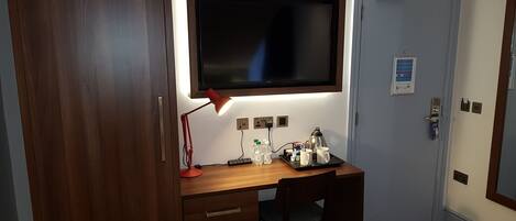 Una camera d'albergo con una scrivania in legno, una sedia, una TV montata sopra la scrivania, un comodino con una lampada e un letto con copriletto decorato.