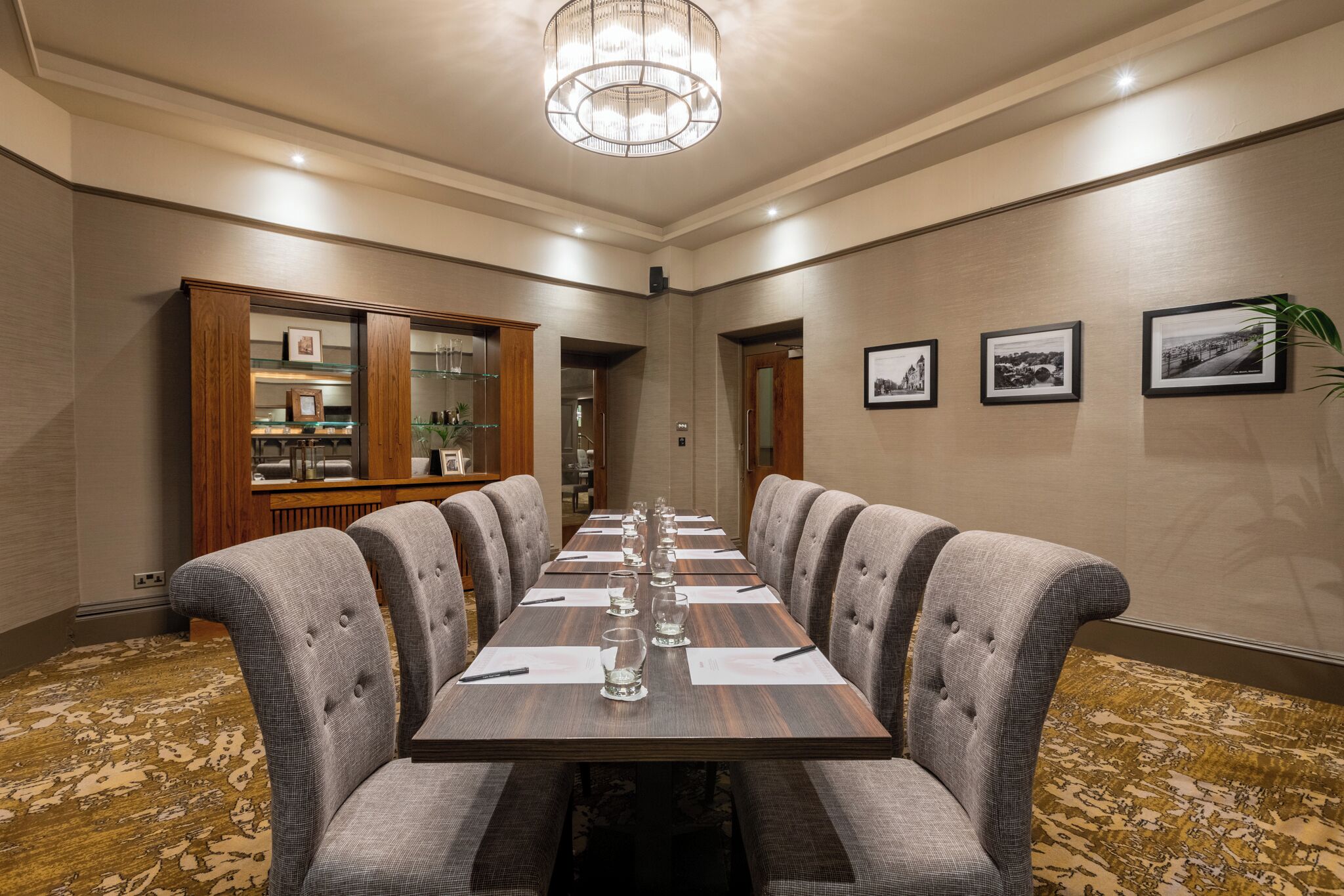 Un salón de conferencias con una mesa larga, sillas tapizadas, un aparador de madera con puertas de cristal y cuadros enmarcados en la pared.