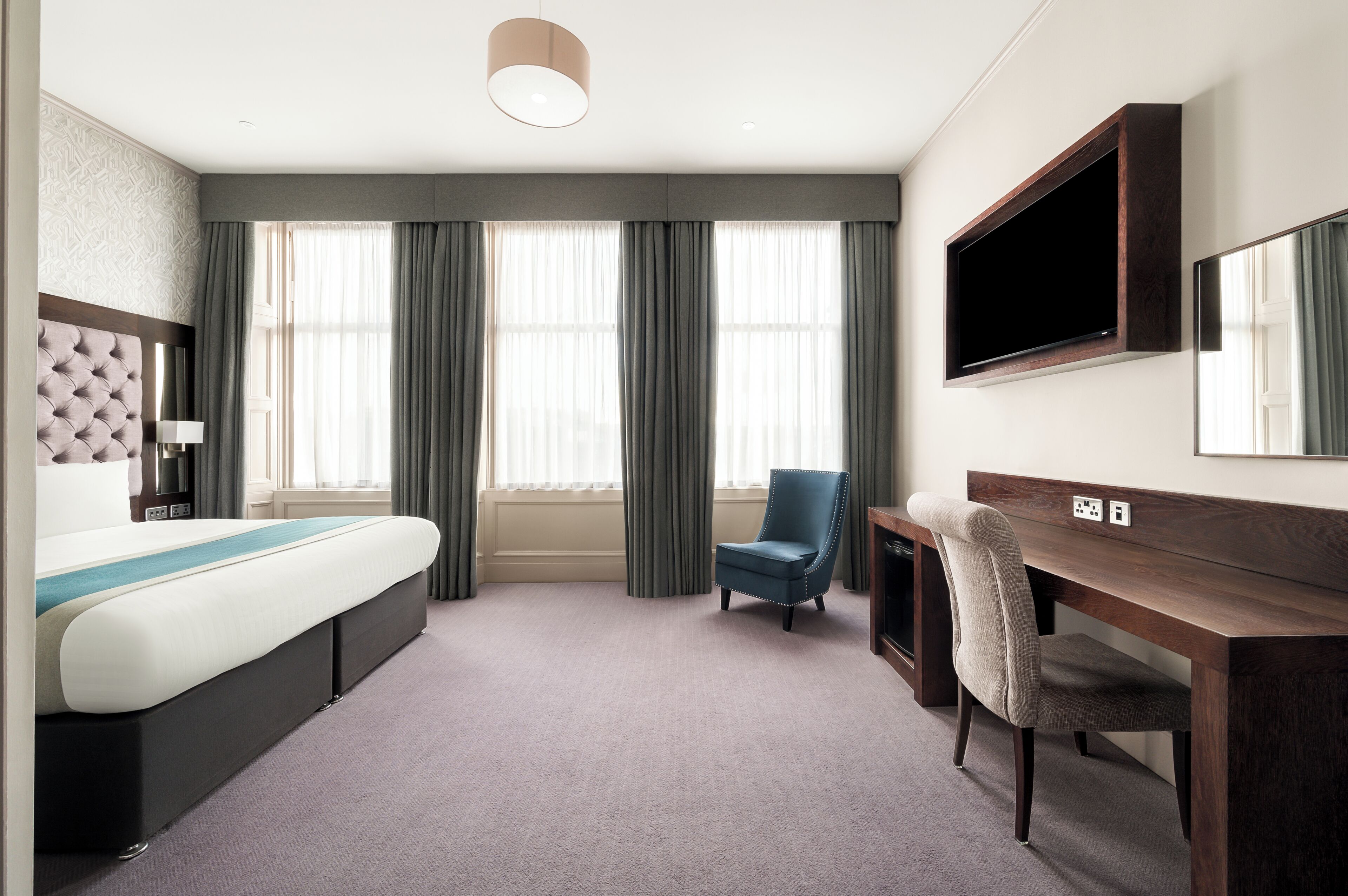 Een hotelkamer met een bed, een bureau, een stoel, een televisie en een groot raam met gordijnen.