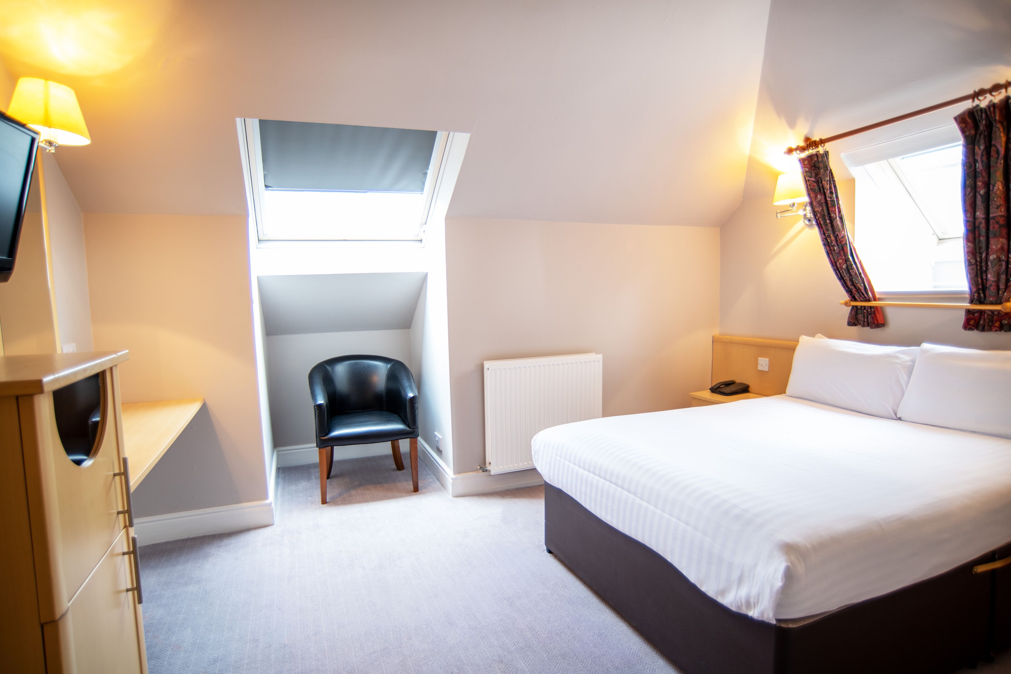 Une chambre à coucher comprenant un lit, une chaise, un bureau, une lampe, une fenêtre avec des rideaux et un radiateur.