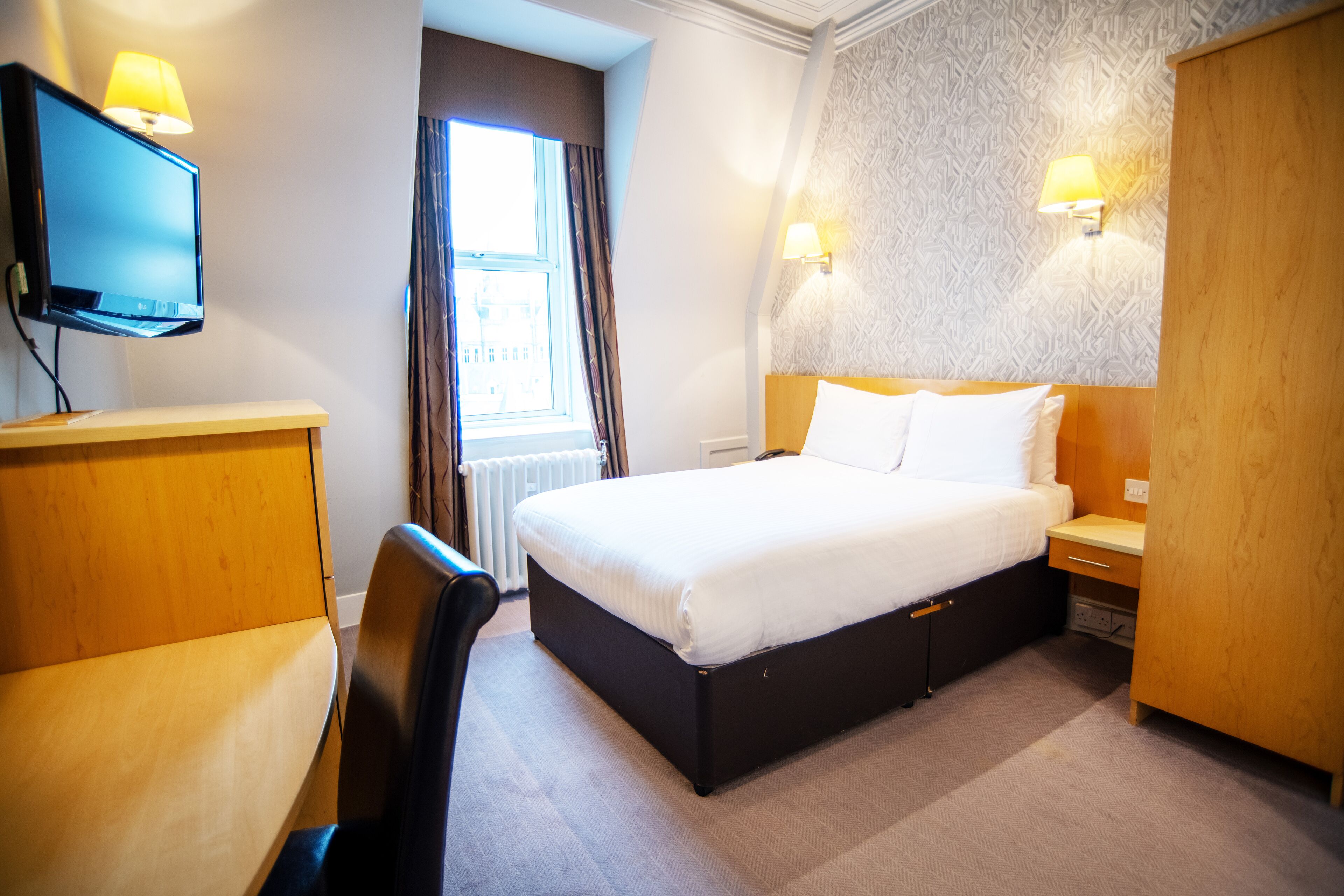 A hotel room with a bed, bedside table, lamp, TV, and a window with curtains.