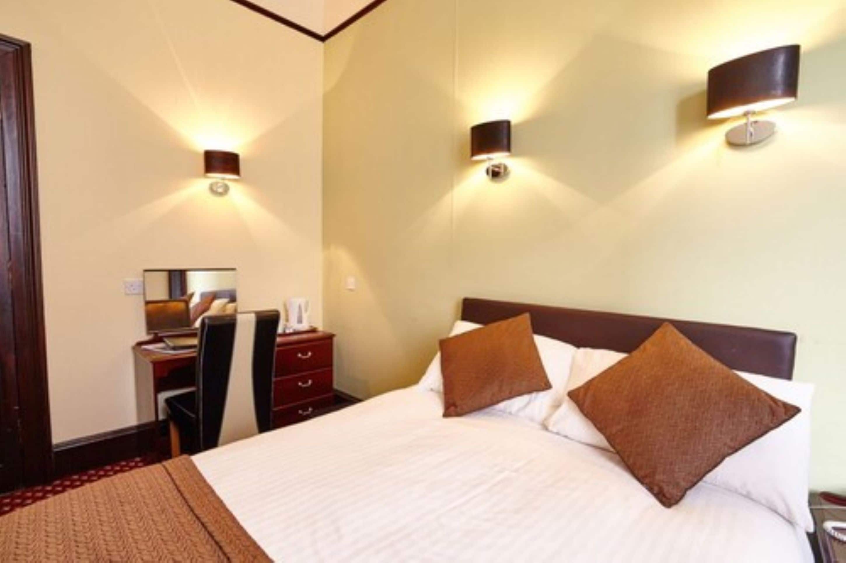 Una habitación de hotel con una cama, un escritorio con una silla y dos lámparas fijadas a la pared.