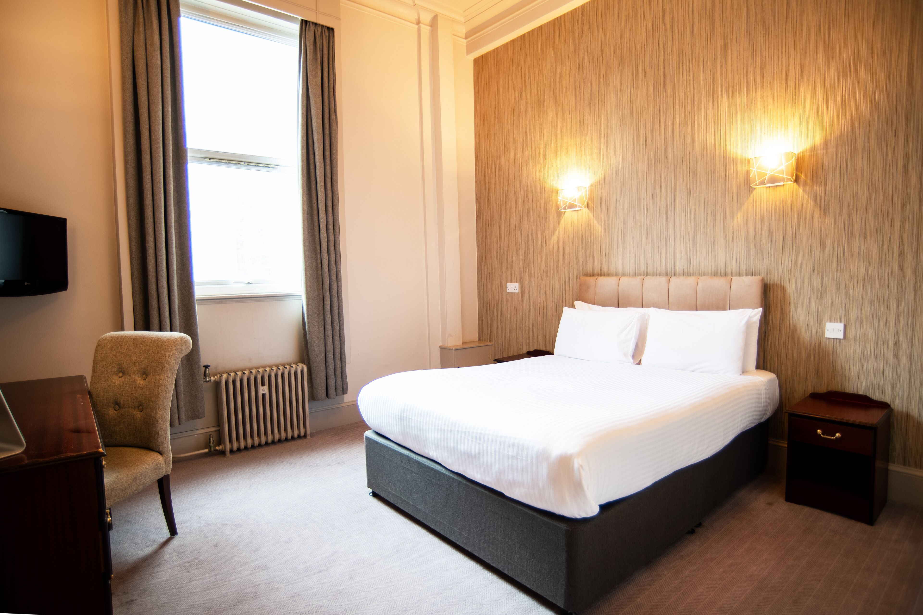 Une chambre d’hôtel avec un grand lit, une chaise, une table de chevet, une télévision et une fenêtre avec des rideaux.