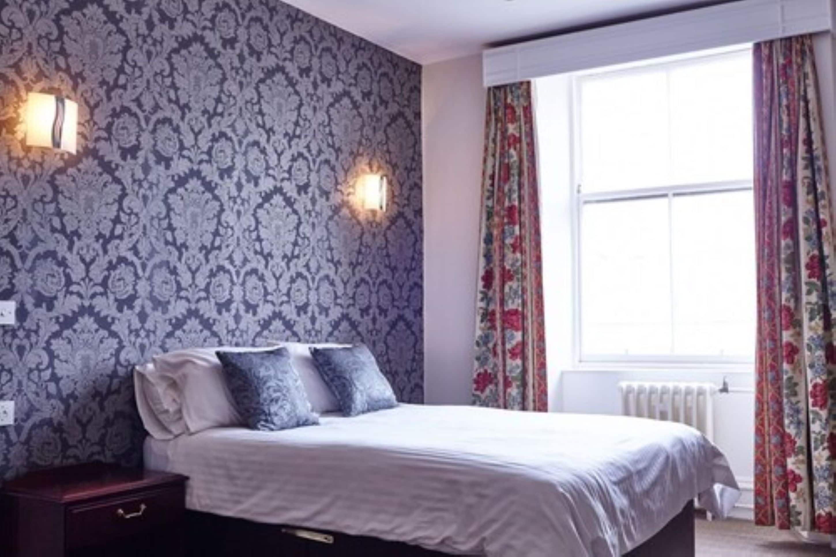 A bedroom with a patterned wallpaper, a bed with white bedding, two wall-mounted lights, and a window with floral curtains.