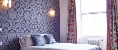 A bedroom with a patterned wallpaper, a bed with white bedding, two wall-mounted lights, and a window with floral curtains.