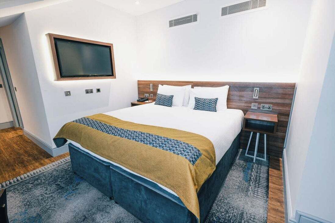 A modern hotel room with a large bed, a flat-screen TV mounted on the wall, and a wooden headboard.