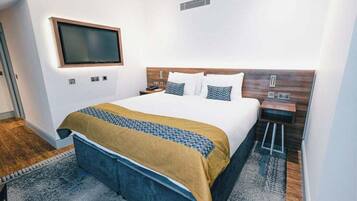 A modern hotel room with a large bed, a flat-screen TV mounted on the wall, and a wooden headboard.