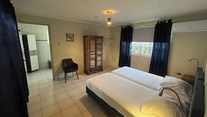 A hotel room with a bed, a chair, a wardrobe, a window with curtains, and a door leading to another room. - Dive Hut - Boutique Apartments (Kralendijk)