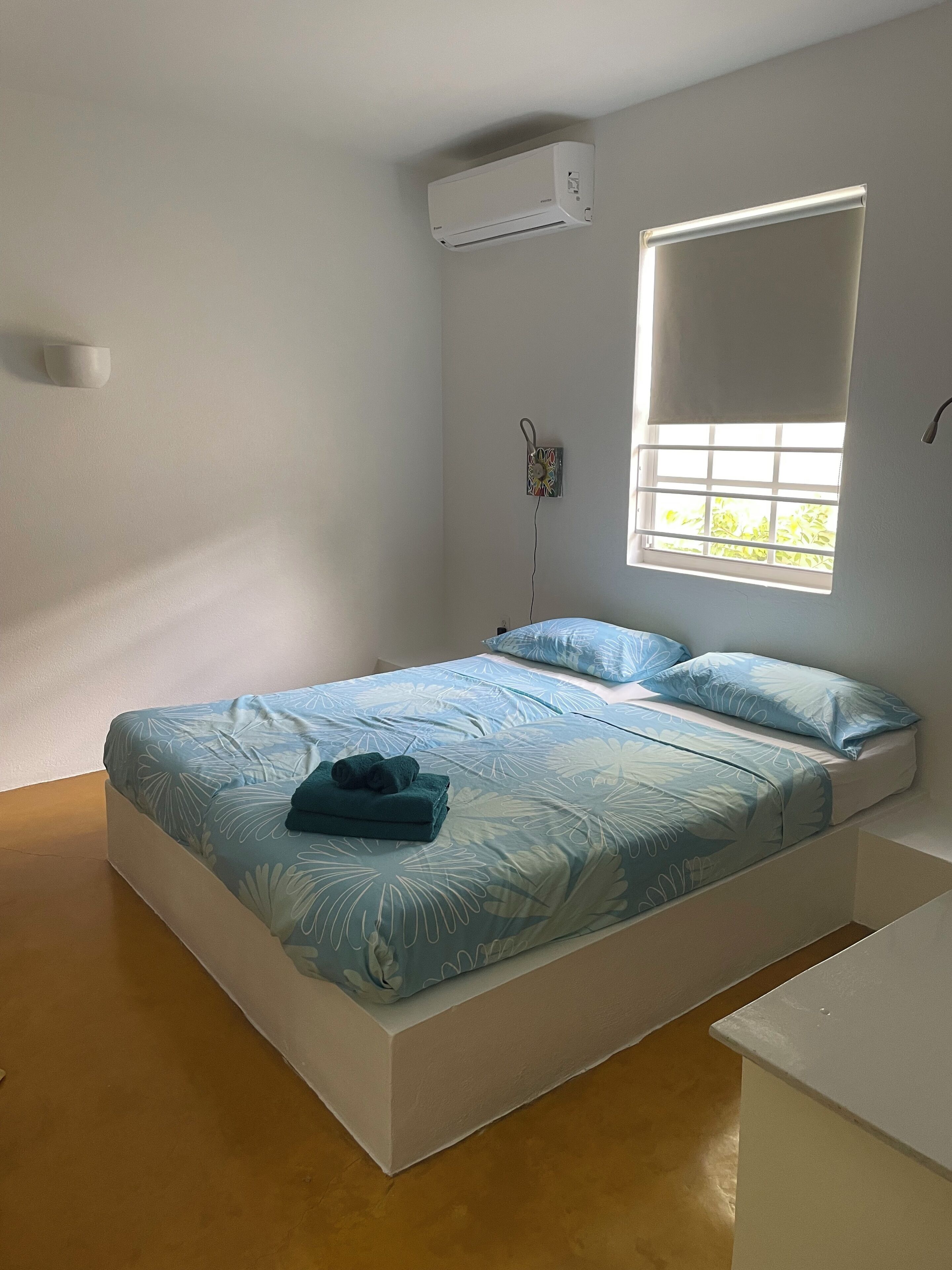 A single bed with a floral bedspread, a bedside table, a window with a view, and an air conditioning unit on the wall.