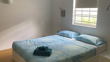 A single bed with a floral bedspread, a bedside table, a window with a view, and an air conditioning unit on the wall.