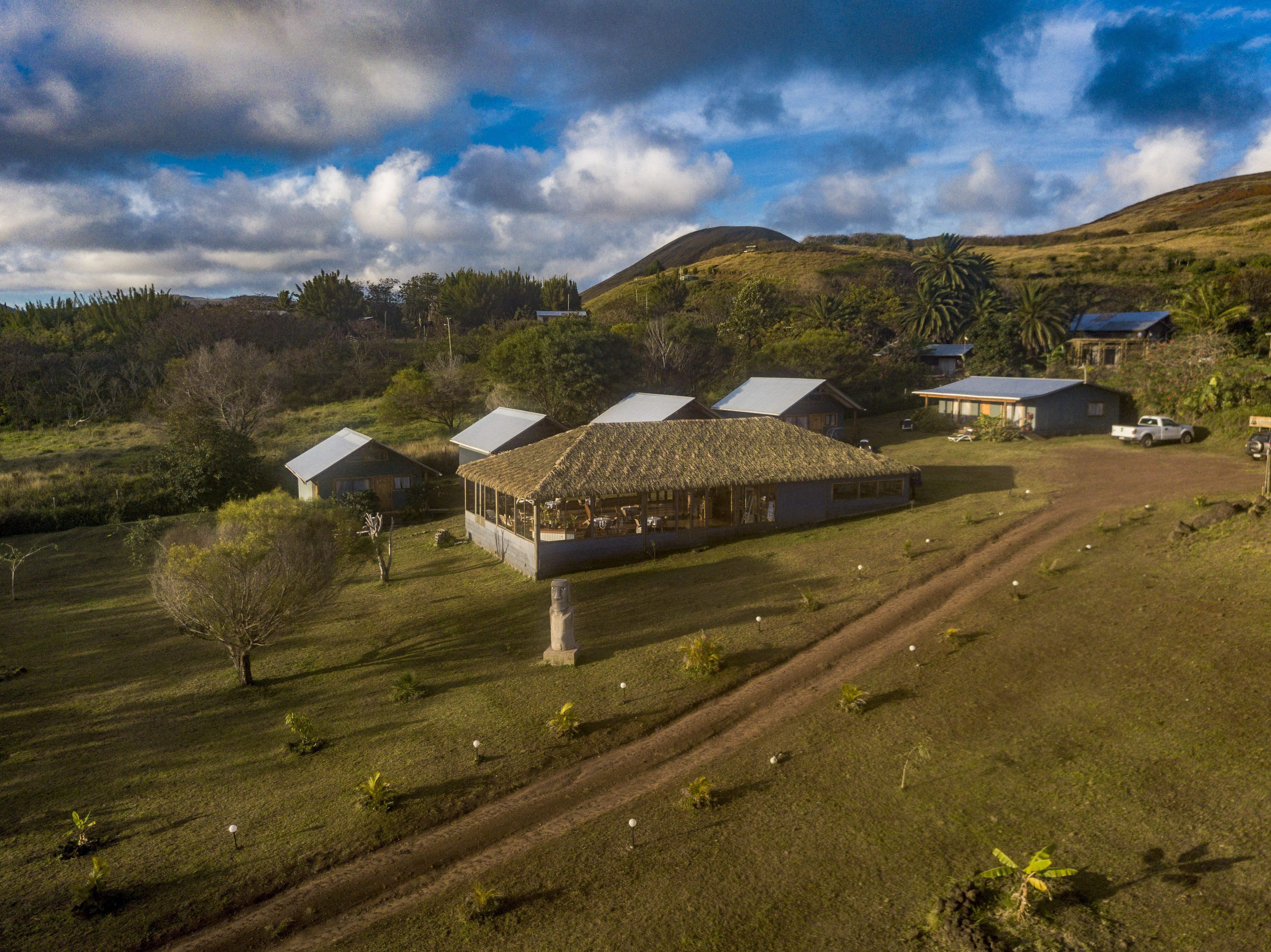 Paisagem rural com várias construções, incluindo uma casa grande com telhado de palha e estruturas menores. Caminhos bem definidos e uma estrada de terra levam às construções.