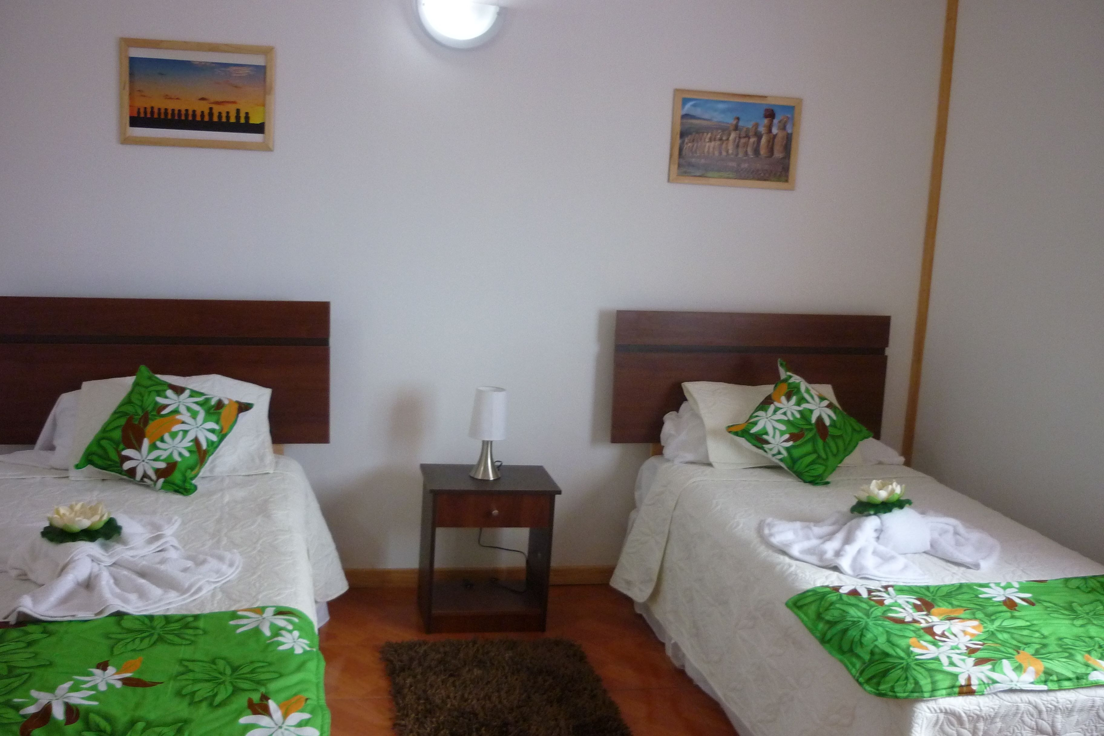 A hotel room with two beds, each with a green floral bedspread, white linens, and matching pillows. There is a nightstand with a lamp between the beds.