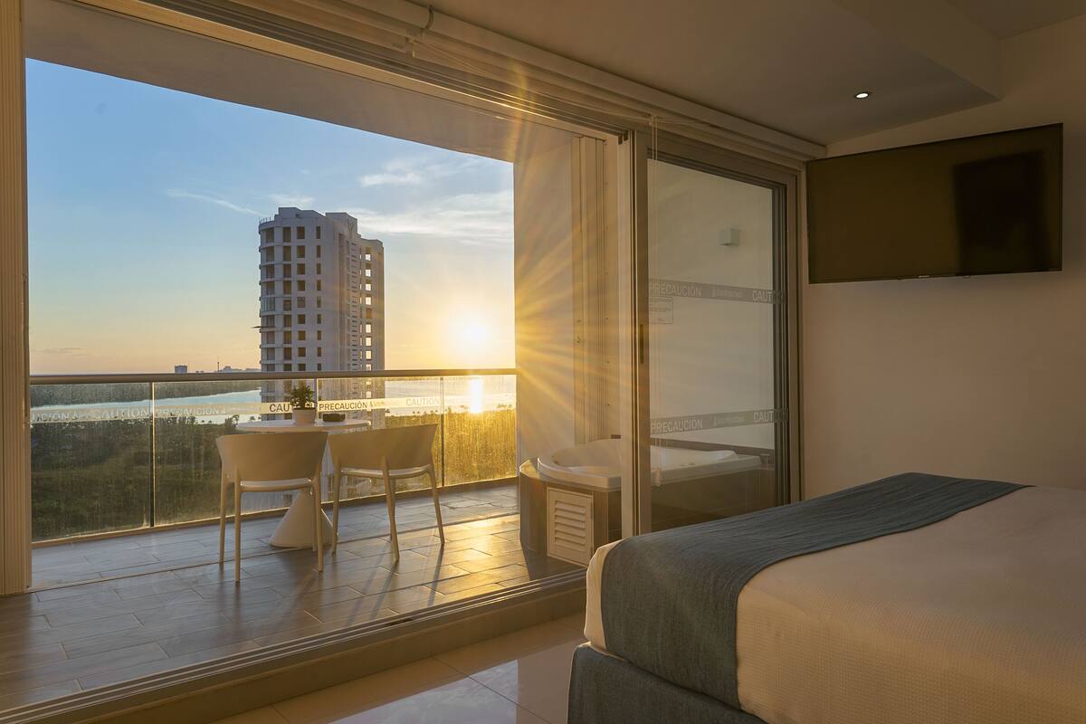 A hotel room with a balcony, a bed, a desk with chairs, and a view of a building and a river.