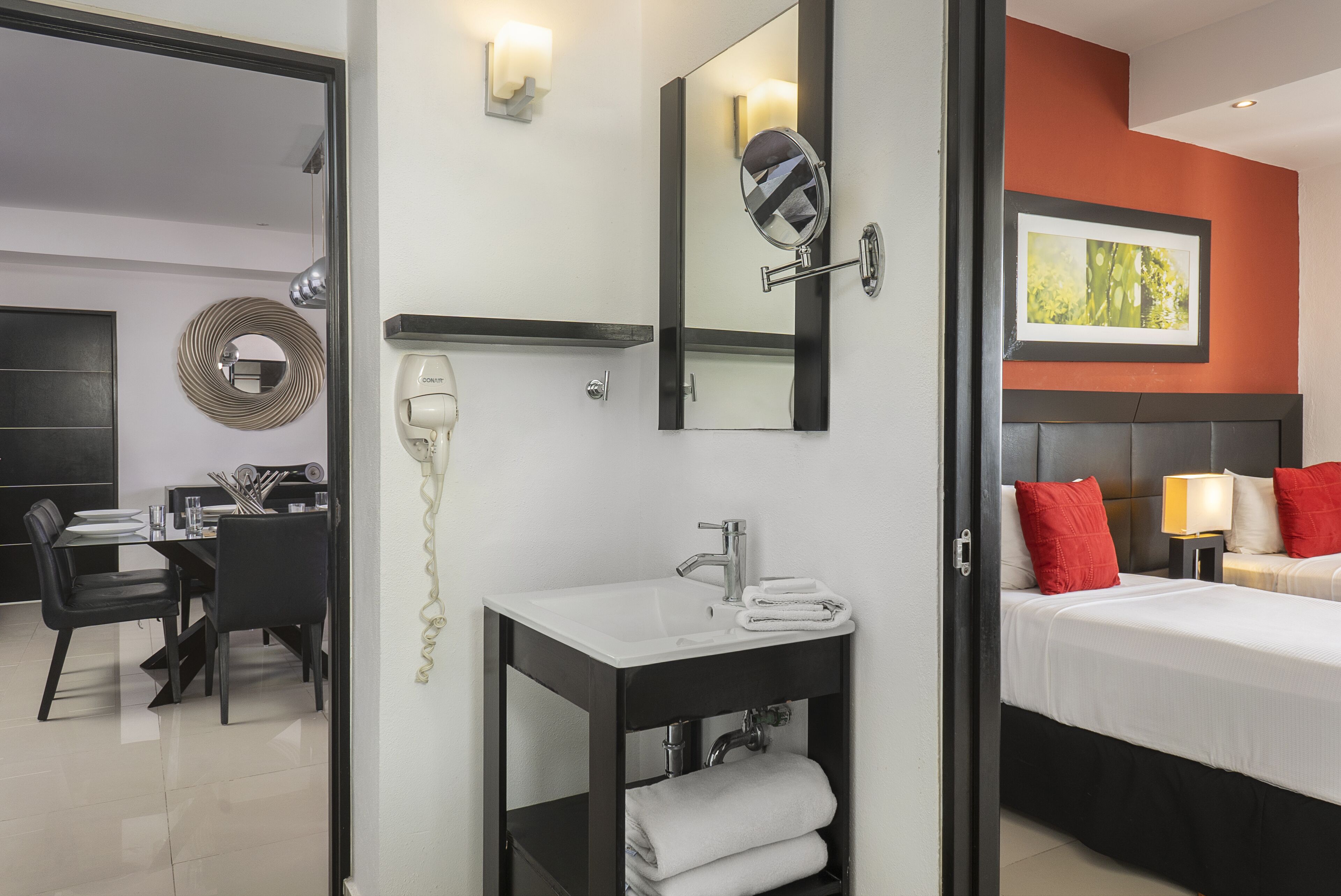 A hotel room with a bed, bedside table, lamp, and a bathroom with a sink, mirror, and hairdryer.