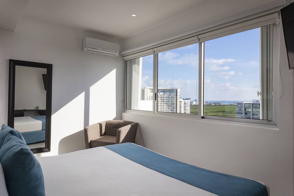 A modern hotel room with a large bed, a chair, a mirror, and a large window offering a city view.