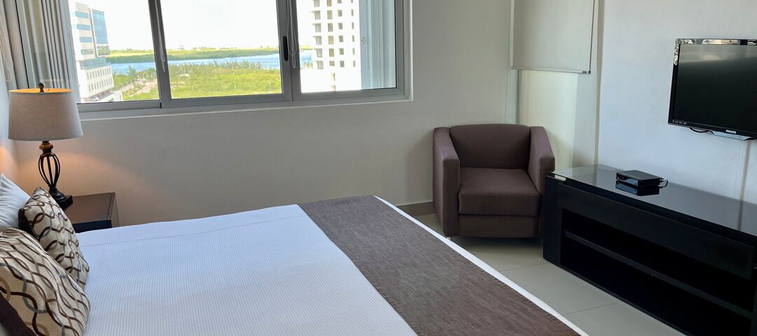 A hotel room with a bed, a chair, a TV, and a window with a view of buildings and water.