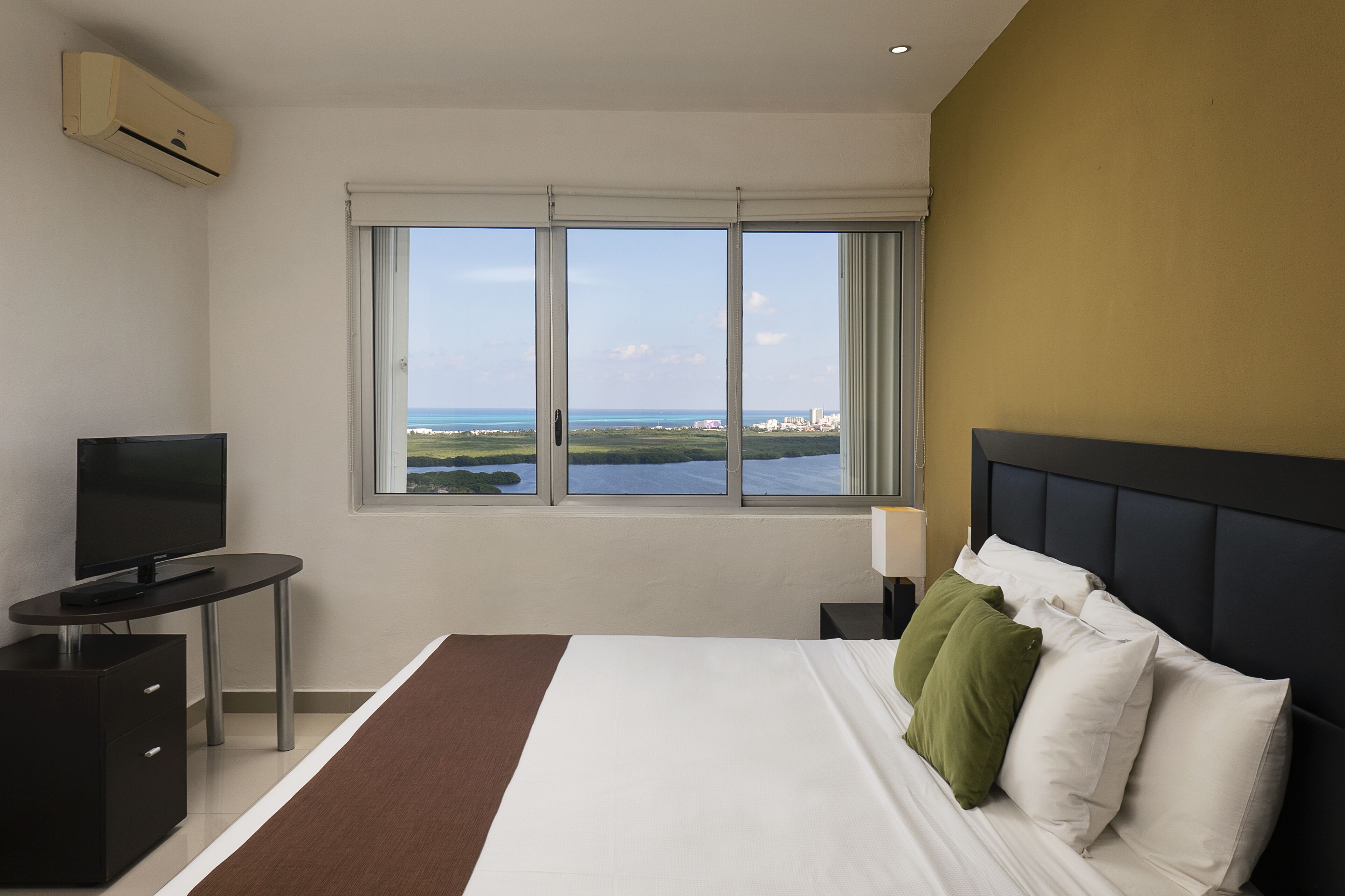 Habitación de hotel con cama, televisor, escritorio y ventana con vista al océano y al horizonte urbano.
