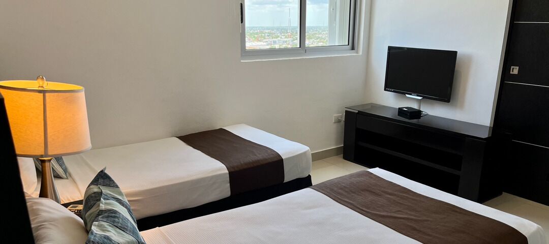 Ett hotellrum med två sängar, en tv på ett stativ, en lampa och ett fönster med utsikt över staden.