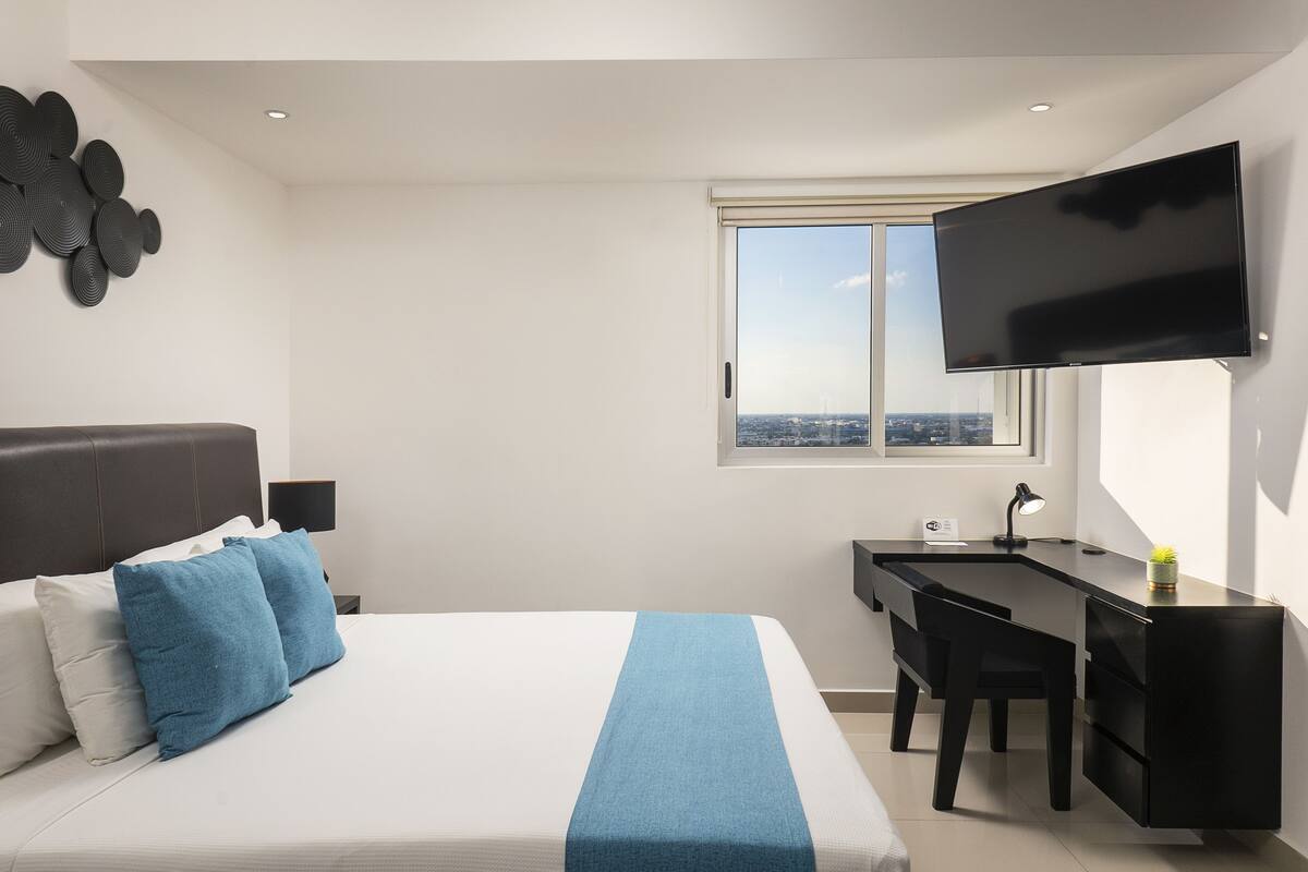 A hotel room with a bed, a desk, a chair, a TV, and a window with a city view.