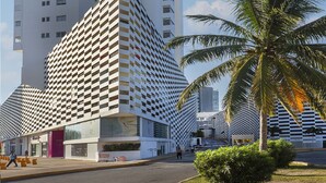 A modern building with a unique checkered facade, a tall structure with a vertical glass section, and a palm tree in the foreground. - Suites Malecon Cancun (Cancun)