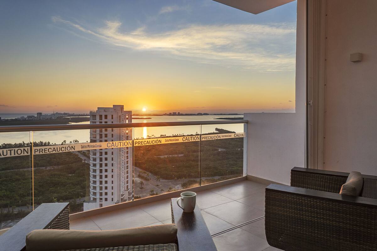 A balcony with a view of a sunset over a body of water, a high-rise building, and a caution tape barrier.