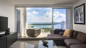 A modern living room with a sofa, a coffee table, and a television on a cabinet. There is a large sliding glass door leading to a balcony with a view of the sea and buildings. - Suites Malecon Cancun (Cancun)