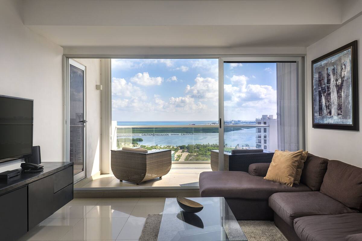 A modern living room with a sofa, a coffee table, and a television on a cabinet. There is a large sliding glass door leading to a balcony with a view of the sea and buildings.