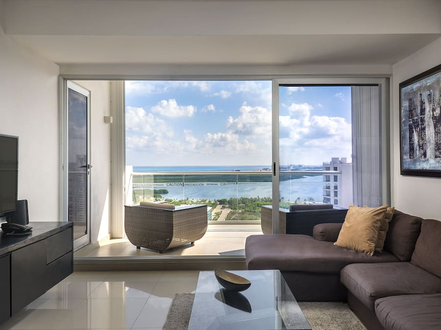Una sala de estar moderna con un sofá, una mesa de centro y un televisor sobre un mueble bajo. Hay un amplio ventanal corredero que da a un balcón con vistas al mar y edificios.