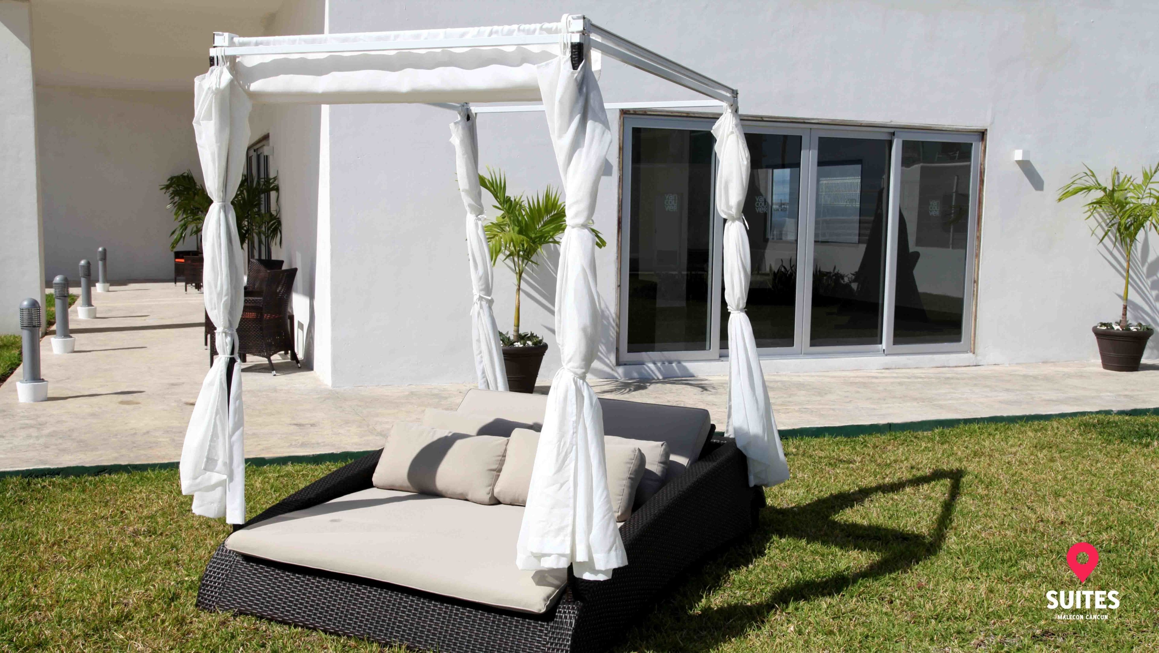 A private outdoor seating area with a canopy bed, a dining table, and chairs, surrounded by a white building and potted plants.