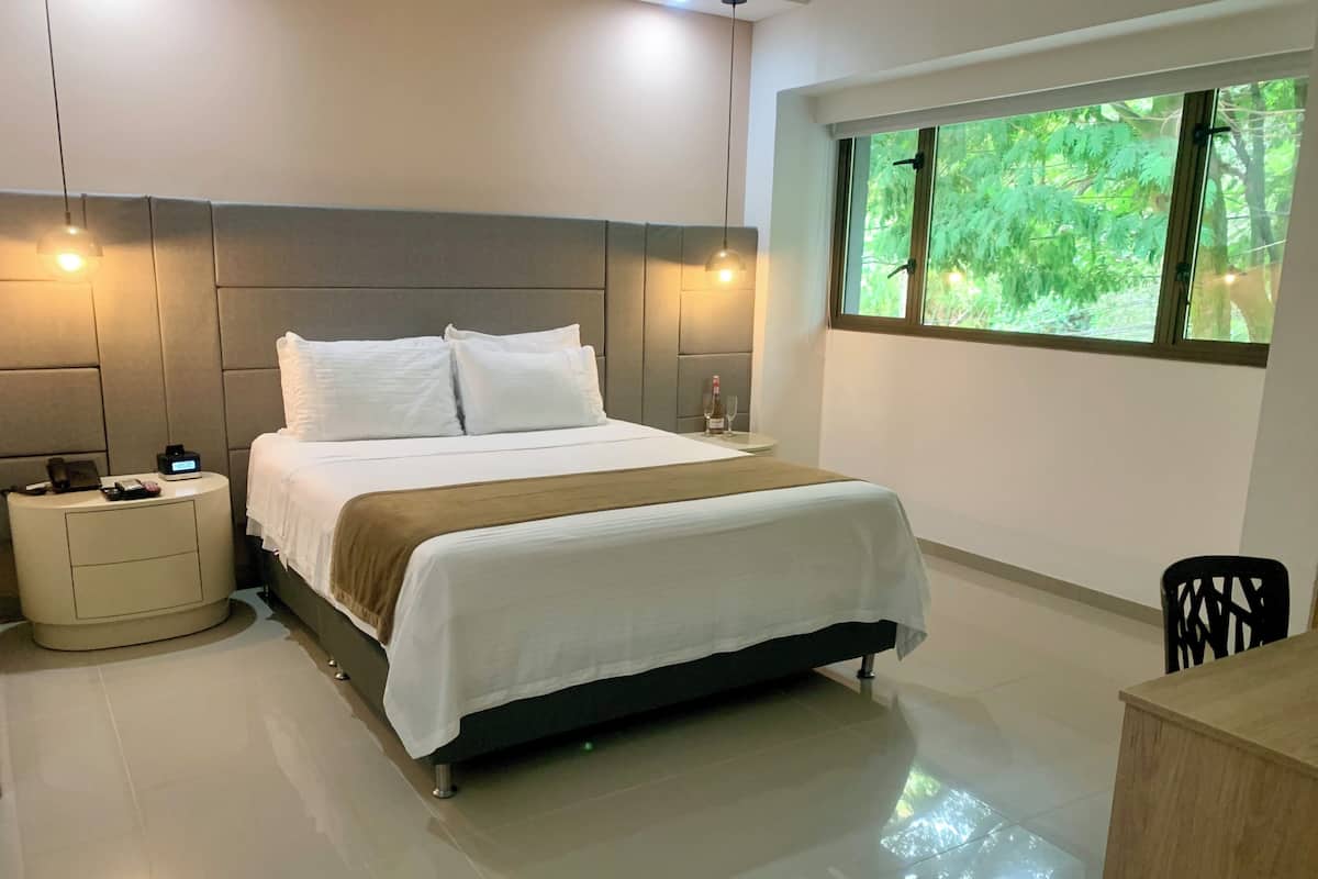 Una habitación de hotel moderna con una cama grande, mesitas de noche, un buró, un ventanal con vistas a zonas verdes y un escritorio con una silla.