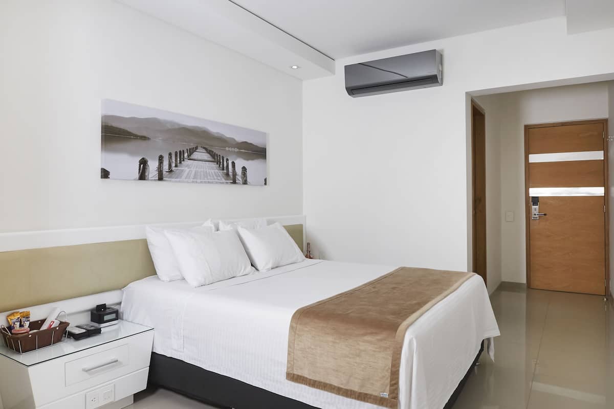Una habitación de hotel con una cama, una mesita de noche con lámpara, aire acondicionado instalado en la pared y una puerta.