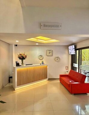 Zona de recepción con mostrador, un reloj, televisor y un sofá rojo.