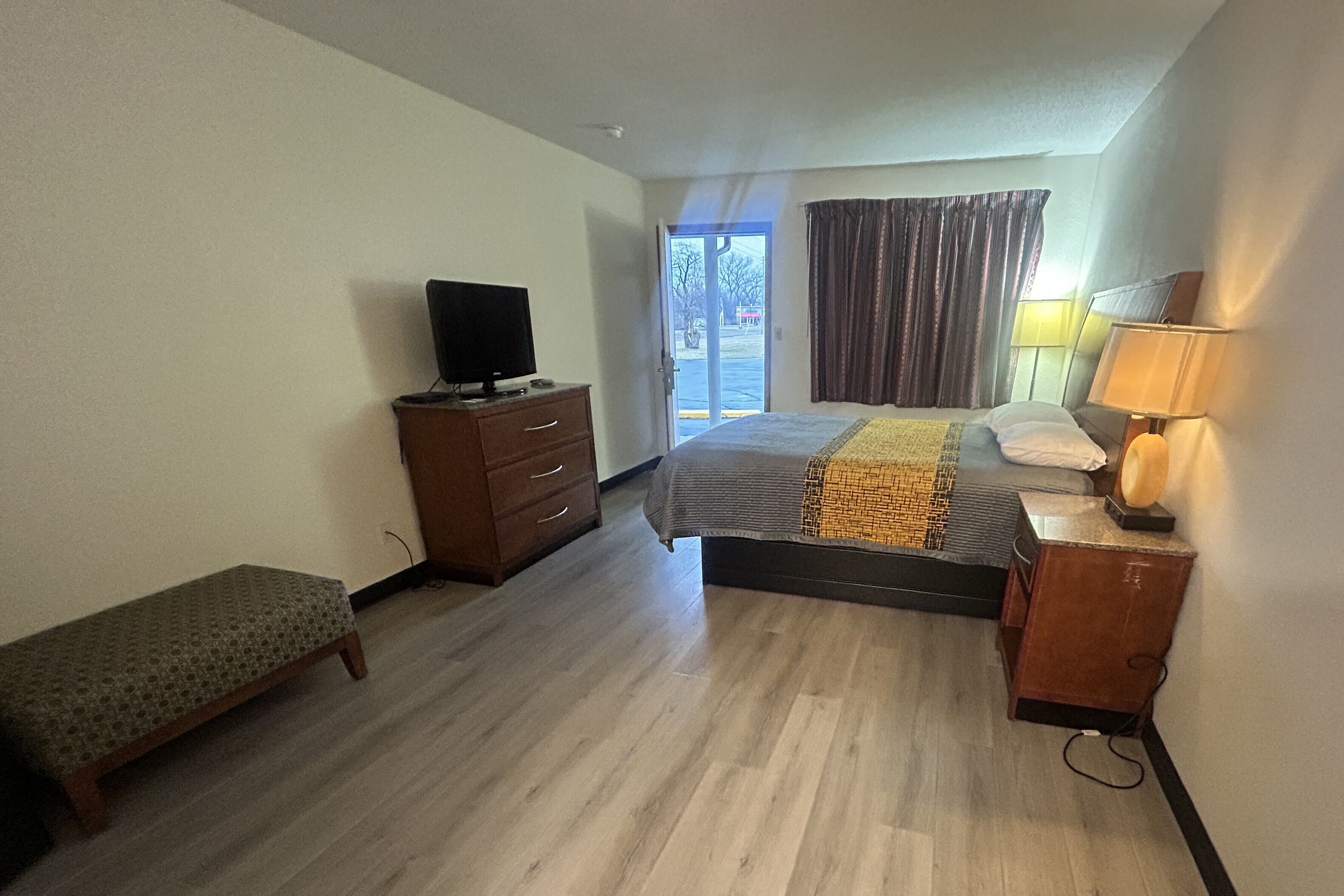 Quarto de hotel com uma cama, um banco, uma cômoda, uma televisão e um abajur.