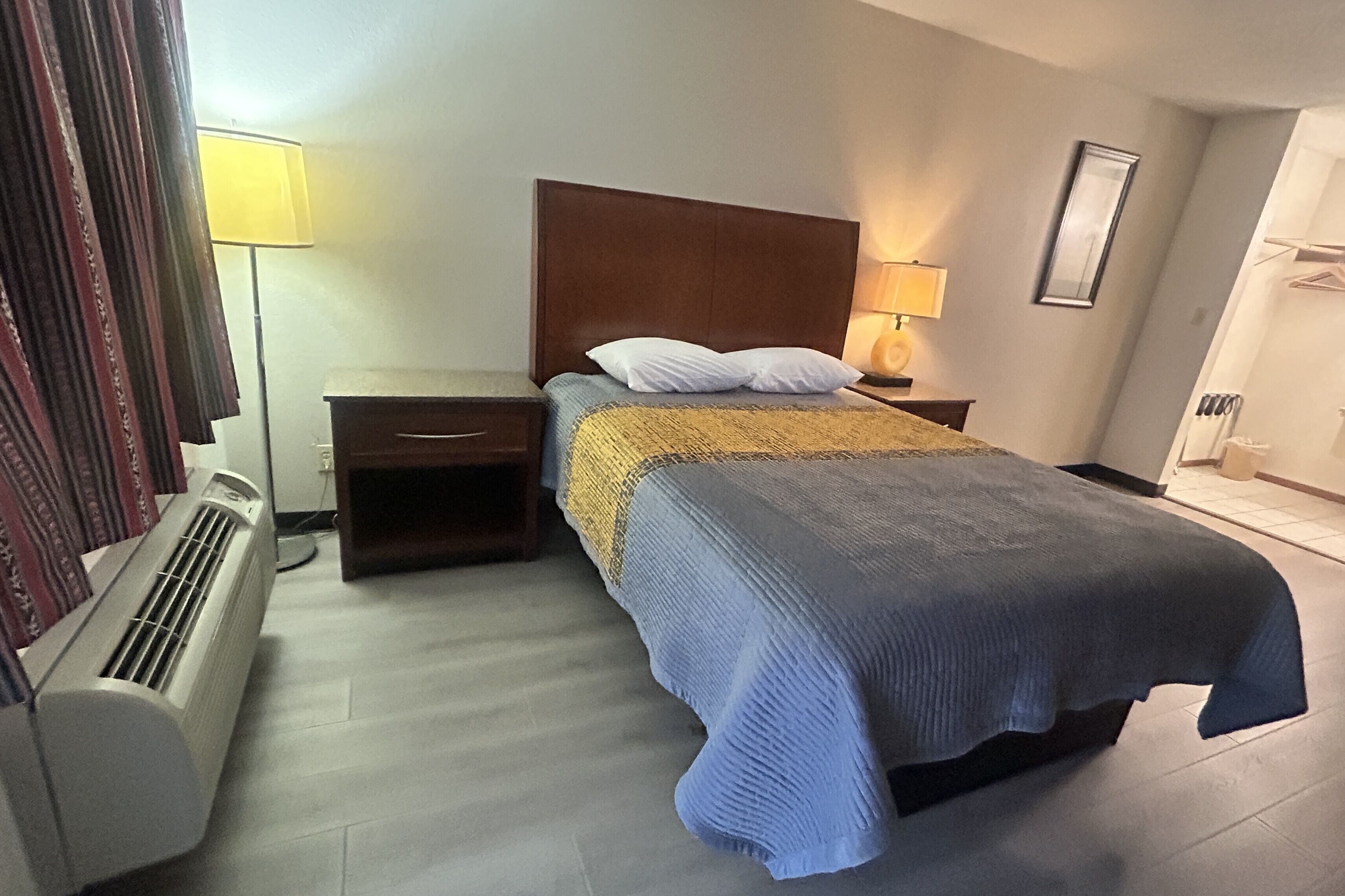 A hotel room with a bed, bedside tables, a lamp, a heater, and a framed picture on the wall.