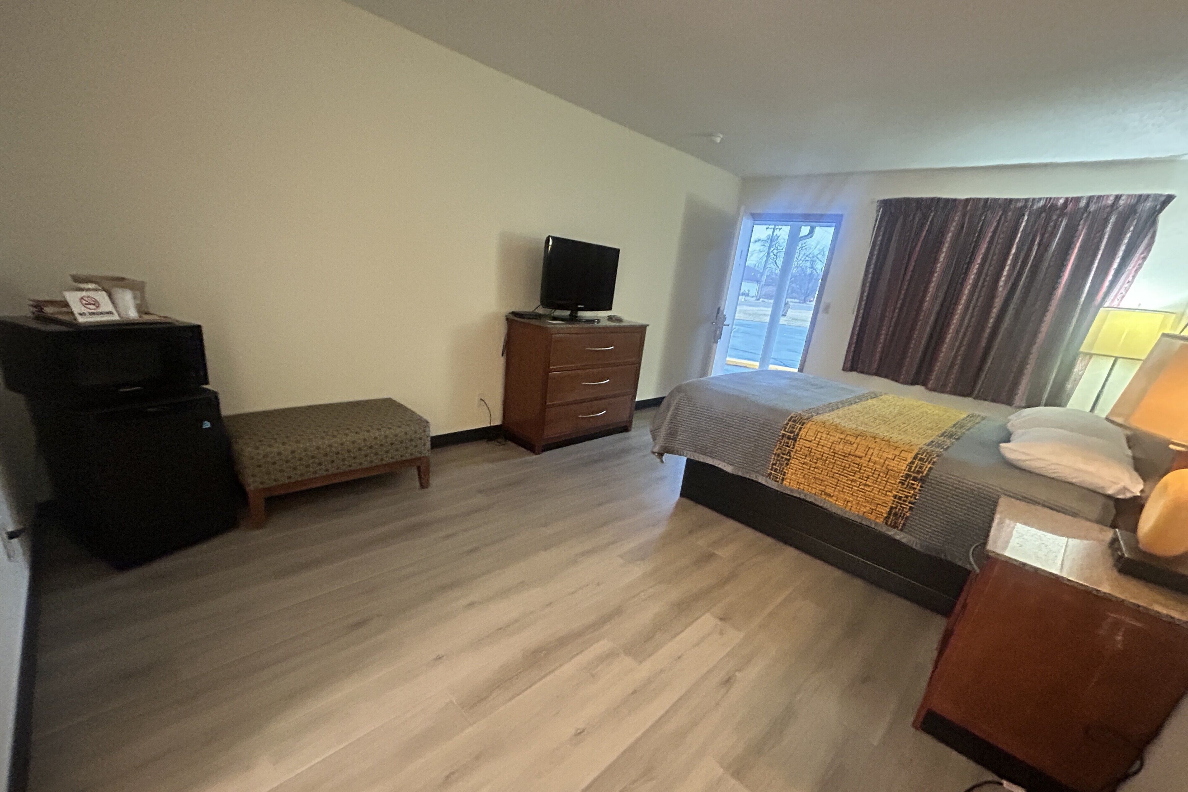 A hotel room with a bed, a television on a dresser, a bench, and a window with curtains.