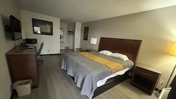 A hotel room with a bed, desk, chair, and a small table with a lamp.