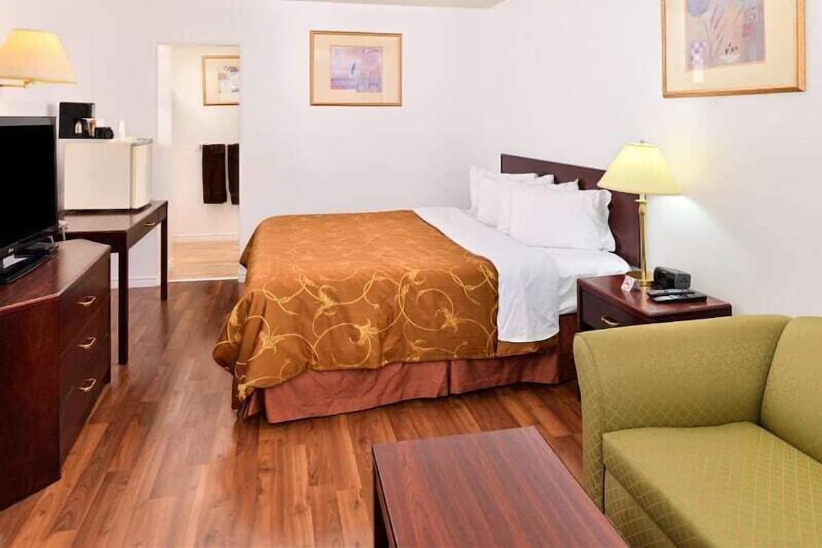 A hotel room with a wooden floor, a bed with a brown and white bedspread, a green armchair, a desk with a lamp, and a television.