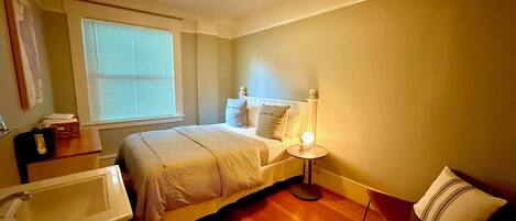 A bedroom with a bed, bedside table, lamp, and a window with blinds.