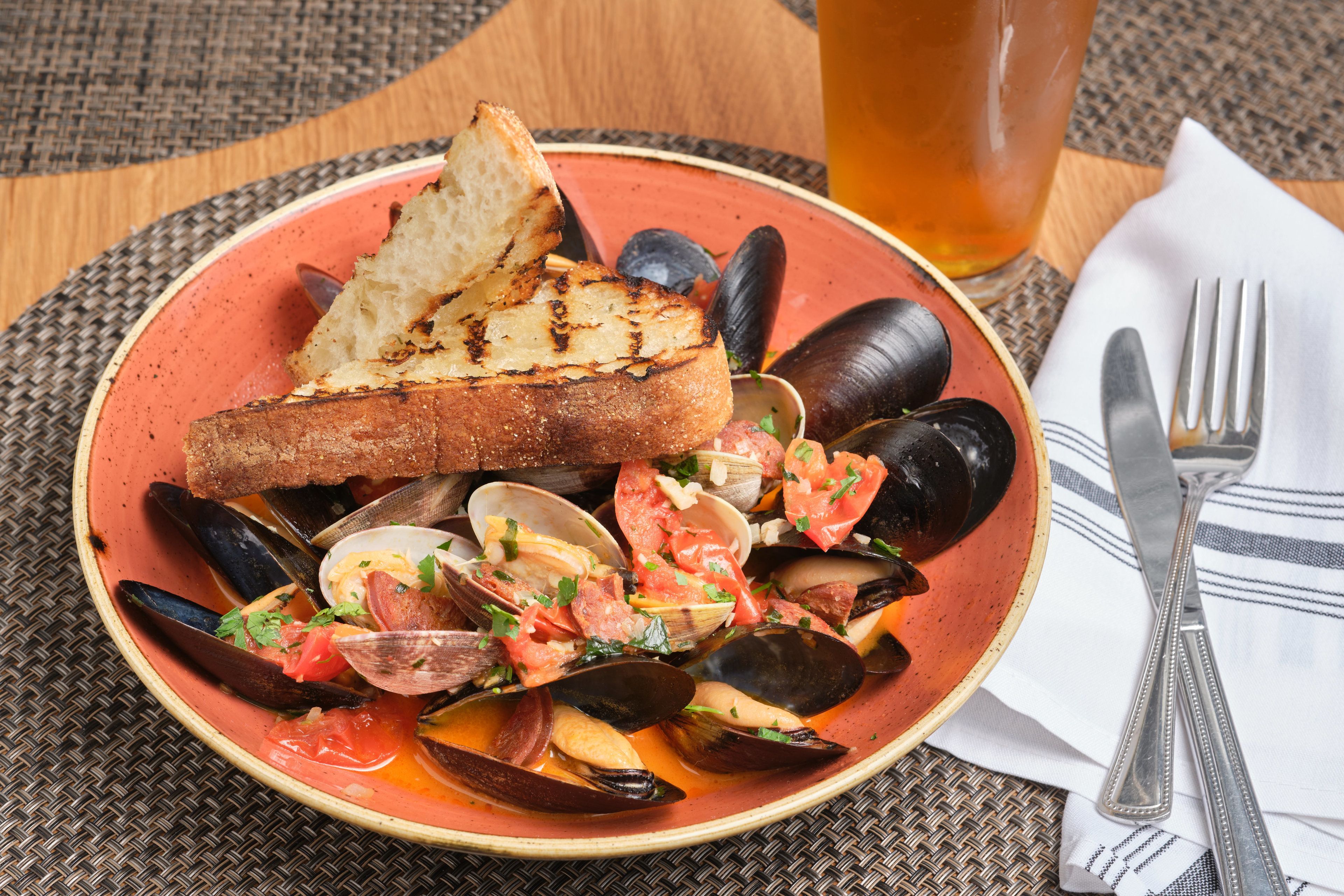 Un plat de fruits de mer composé de moules, de palourdes et d’une tranche de pain grillé, présenté sur une assiette, accompagné d’un verre de bière et de couverts sur une table.