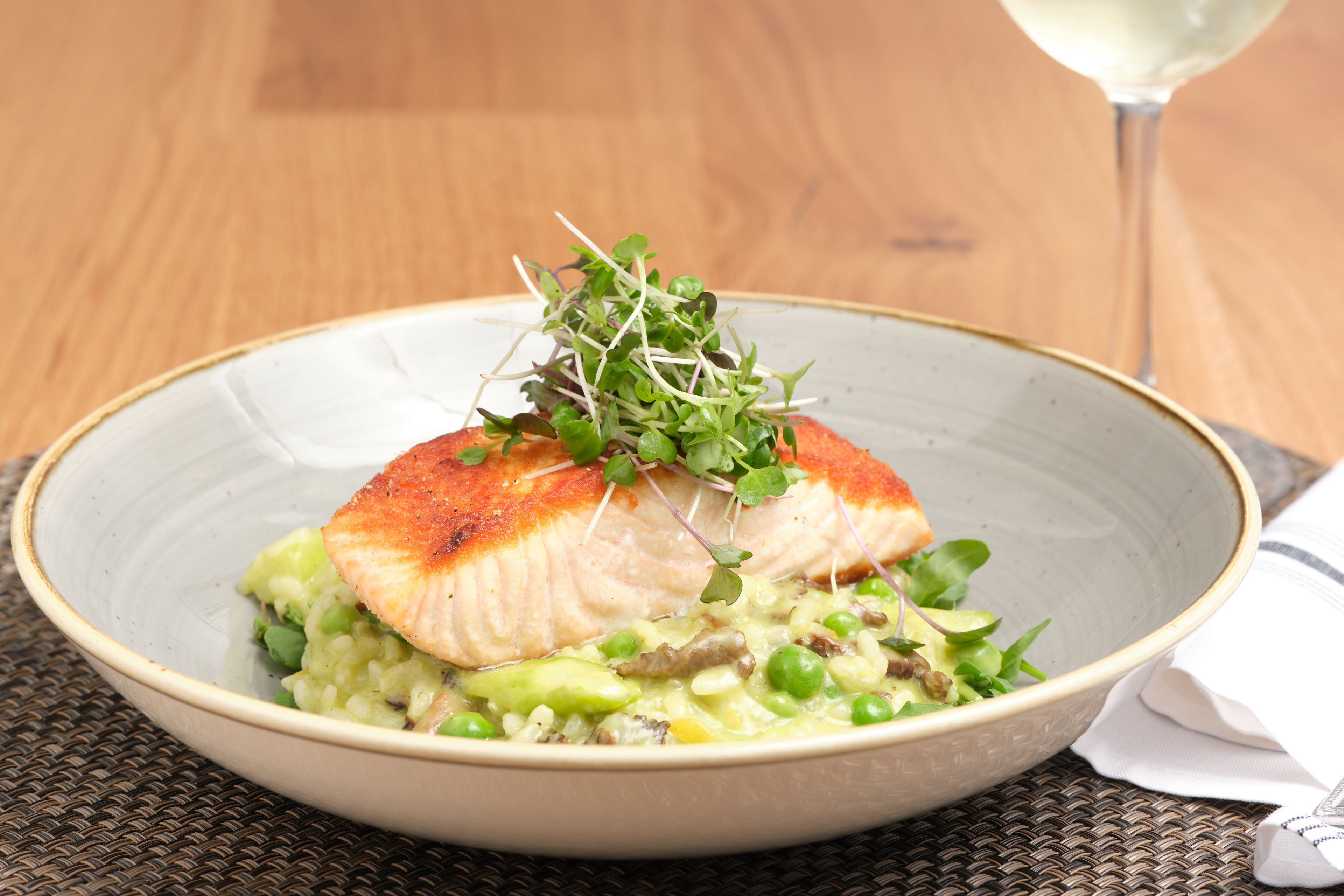 A bowl of food with a piece of fish on top, garnished with greens, and a glass of white wine in the background.