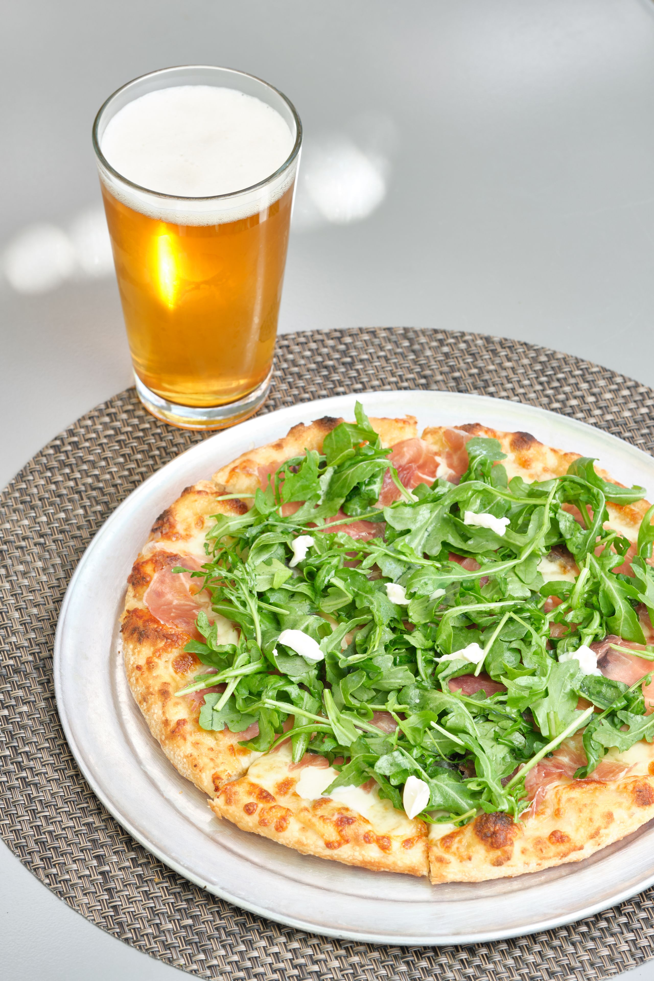 A pizza with arugula and prosciutto on a white plate, accompanied by a glass of beer on a woven placemat.