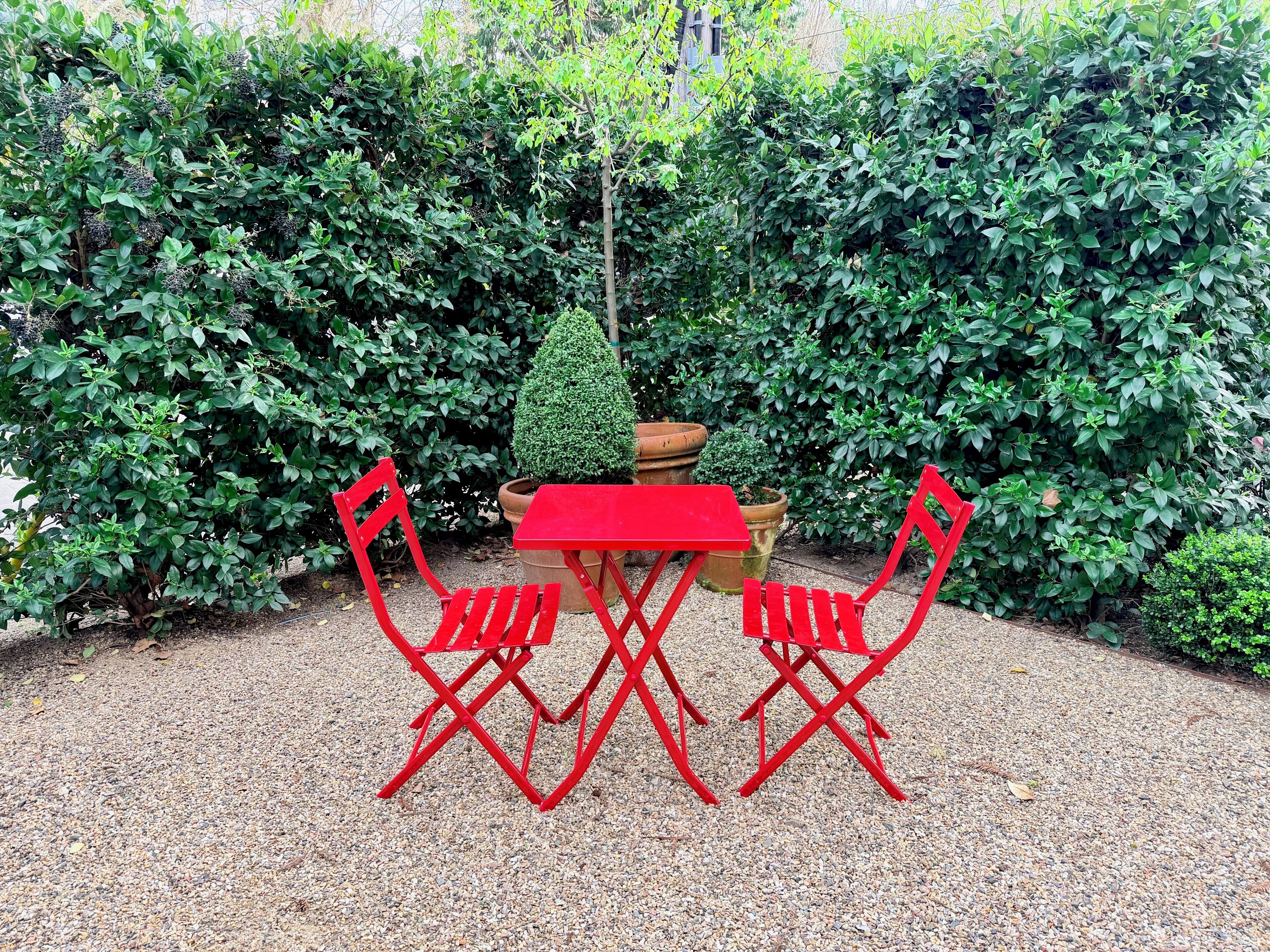 A red outdoor table and chairs set in a garden with dense green foliage in the background.